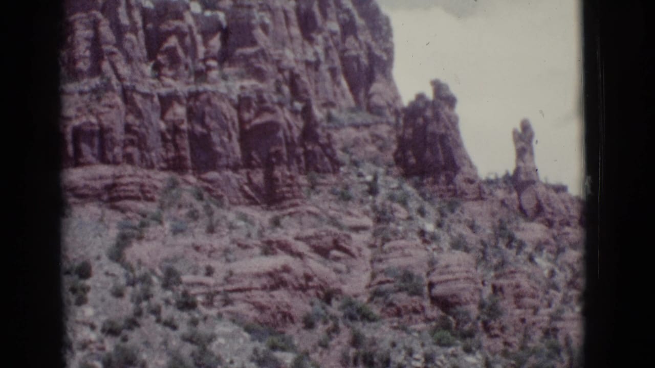 Rocky red cliff face with scrub vegetation and a narrow stone spire under a pale sky.
