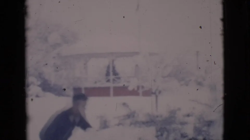A handheld 8mm frame shows the man shoveling snow in Clarkdale, Arizona, with a snow-covered home and trees behind him.