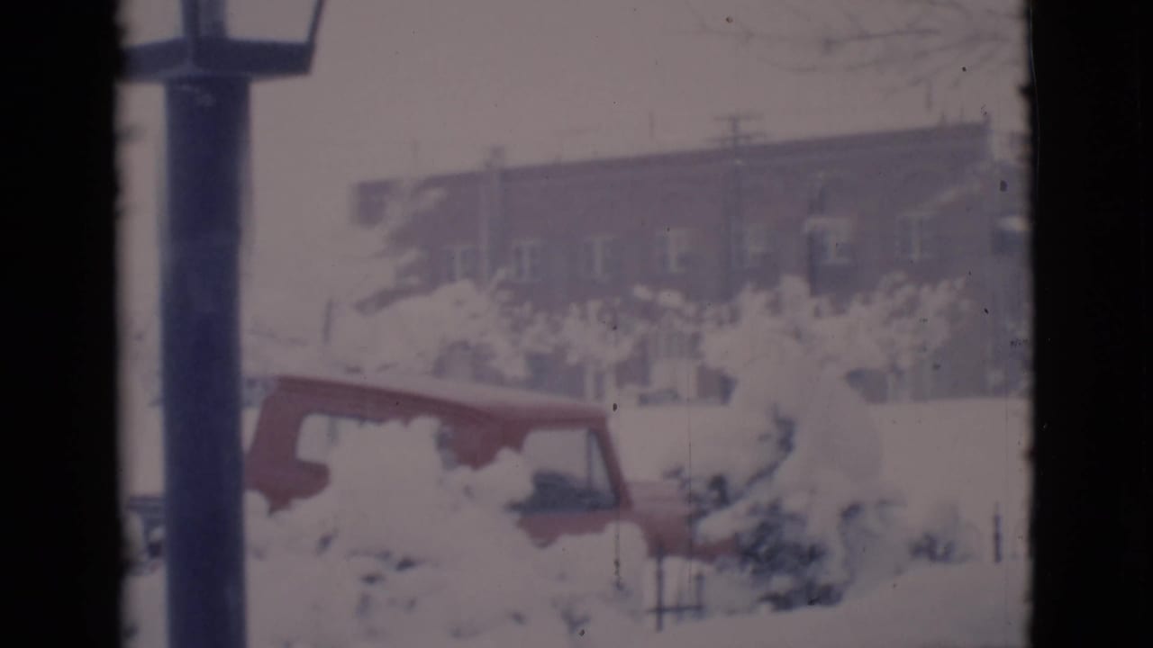 Snow-covered yard with a red car partly buried and a brick building beyond.
