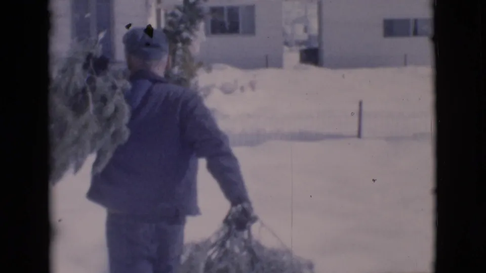 Seen from behind, the man carries or drags a Christmas tree across deep snow near a light-colored house in the 1966 home movie.