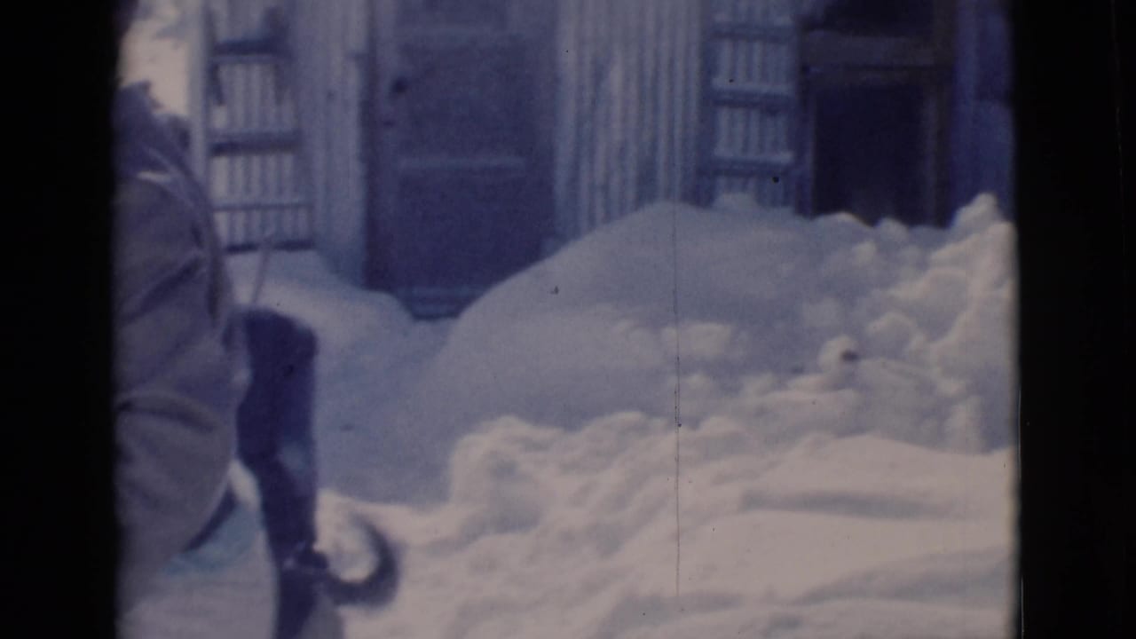Person in winter clothing stands near deep snow beside a corrugated building.