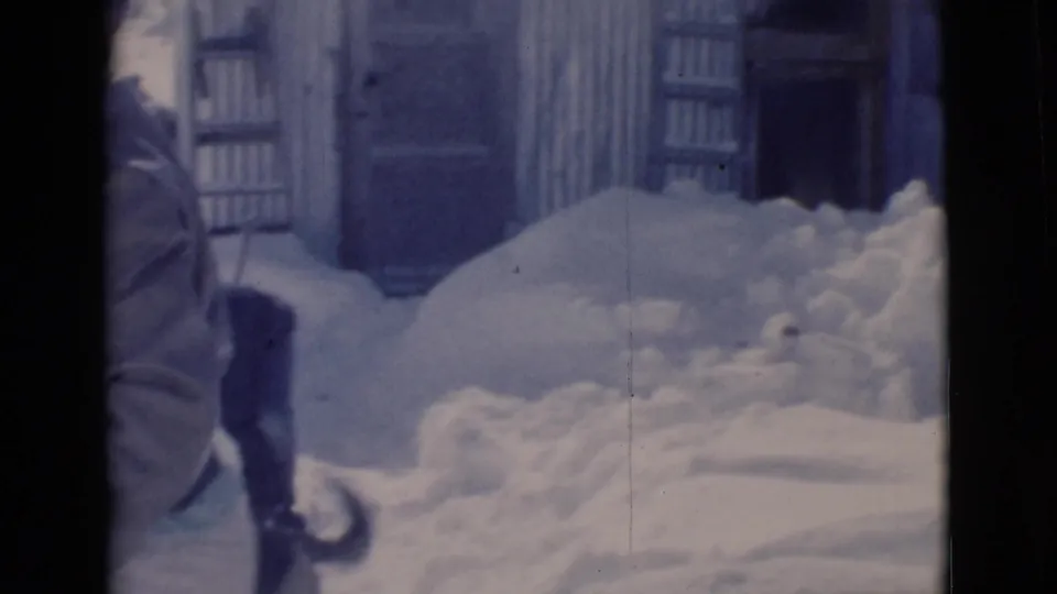 1966 Clarkdale, Arizona home-movie frame with a bundled person beside heavy snowdrifts near a corrugated outbuilding.