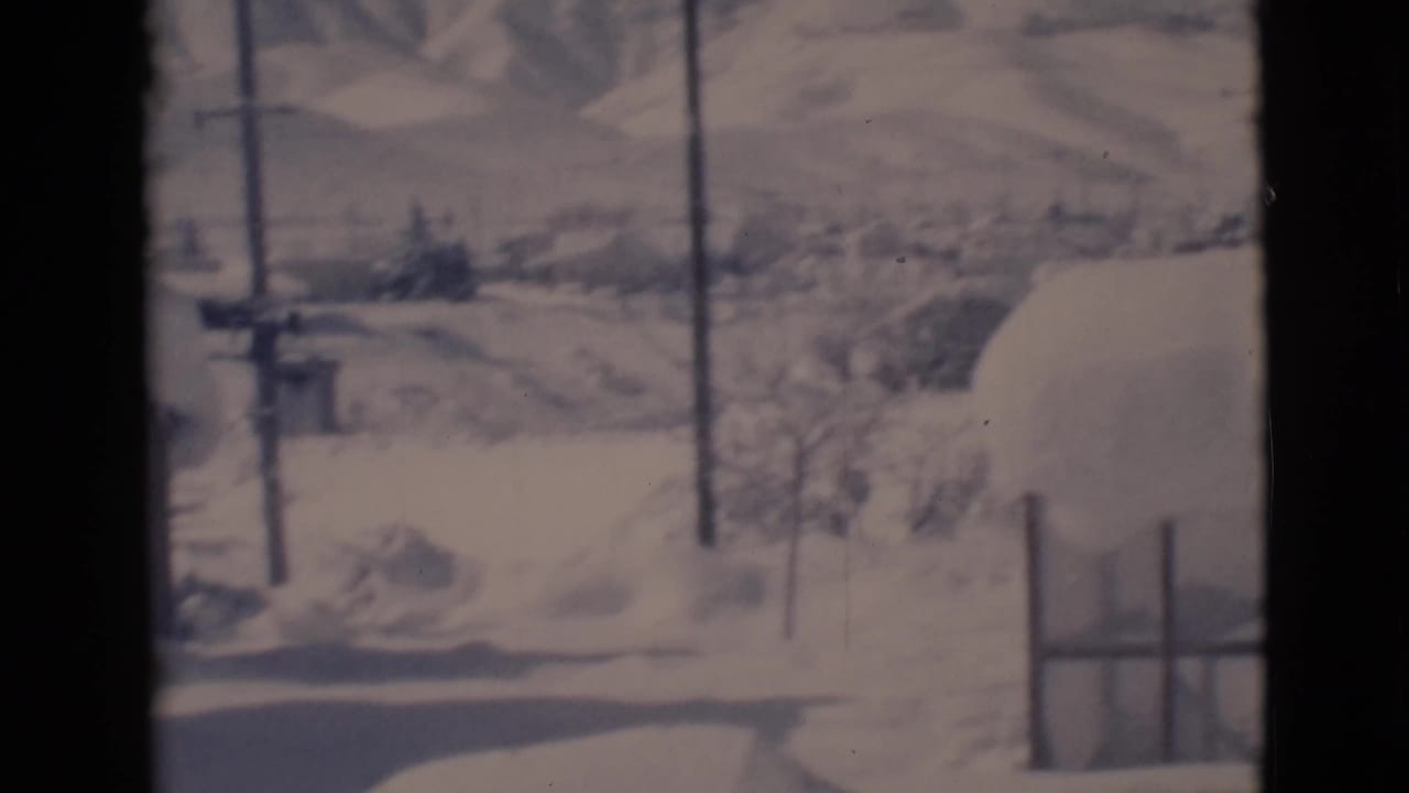 Snowy roadside with utility poles, low hills, fencing, and buried ground.