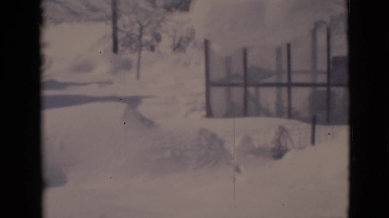 Snow-covered yard with fence posts, bare trees, and a house edge in the distance.