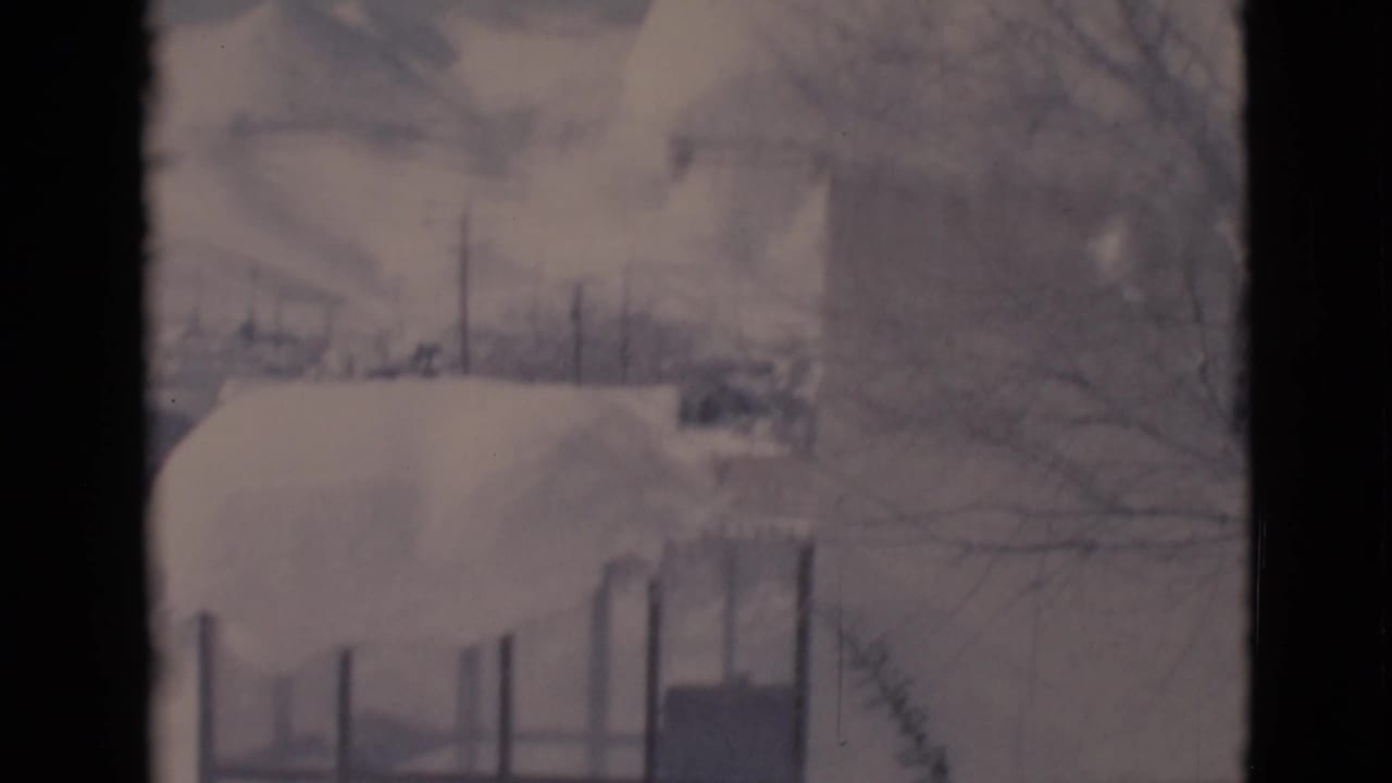 Snow-covered roof or structure with fence posts and bare trees behind it.