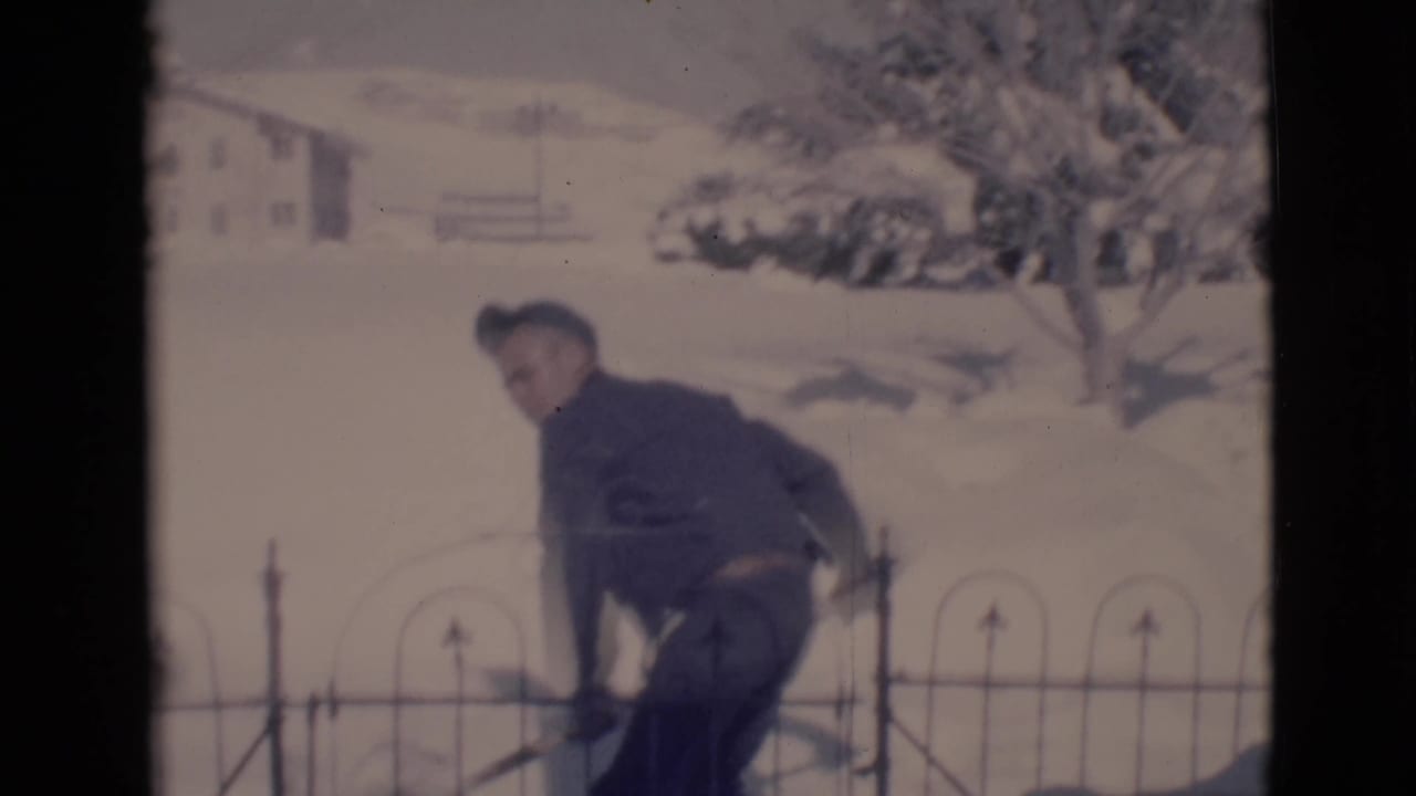 Youth in a dark jacket bends with a snow shovel beside a black metal fence in a snowy yard.