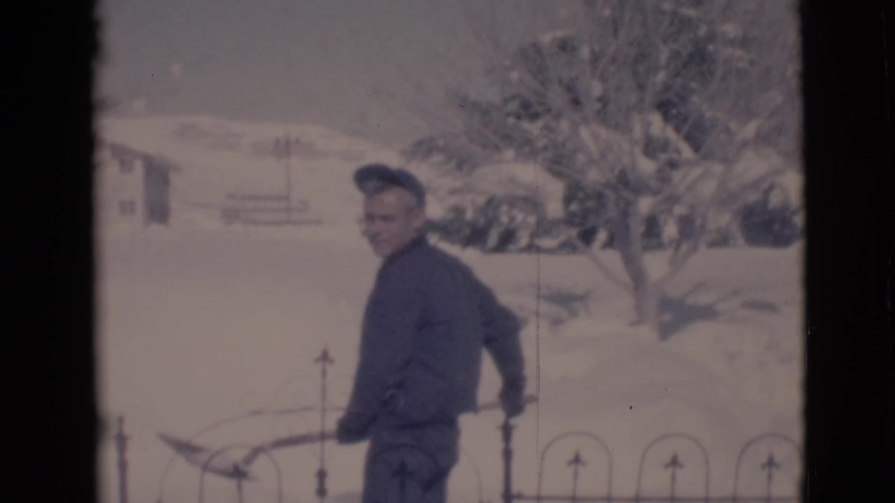 Youth stands in profile holding a snow shovel near a black fence with snow-covered trees behind.
