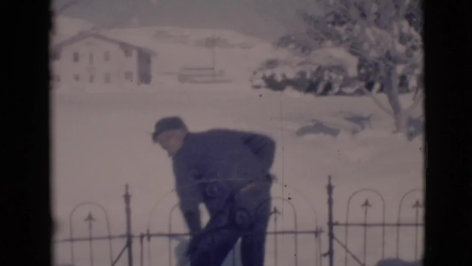A youth bends forward to gather snow with a shovel in the 1966 Clarkdale yard, framed by fence rails and a snowy house beyond.