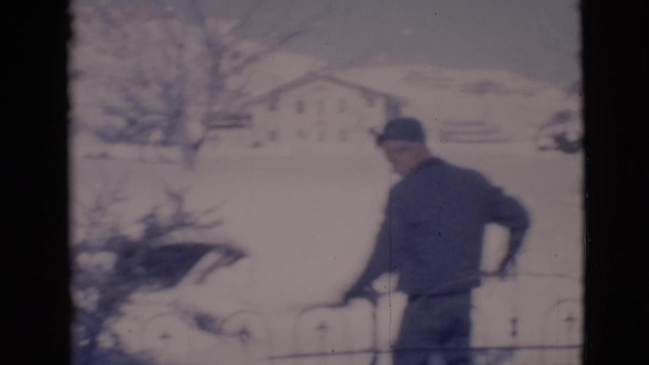 Man in a dark coat and cap shovels snow beside an iron fence in a snowy yard.