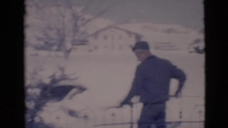 In 1966 Clarkdale, Arizona footage, a man in a dark coat and cap shovels snow near an iron fence with a house behind him.