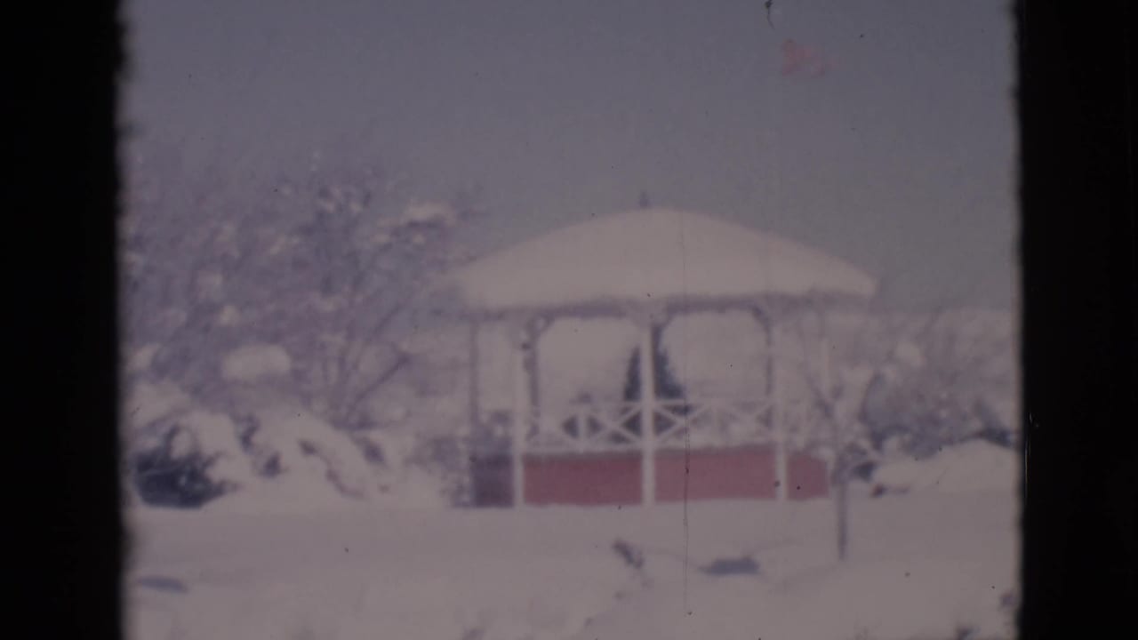 White gazebo with red base surrounded by snow-covered trees and shrubs.