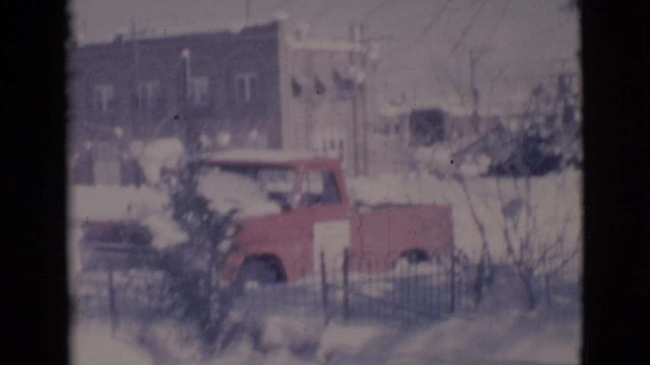 Red pickup truck parked by snowbanks with buildings and fencing behind it.