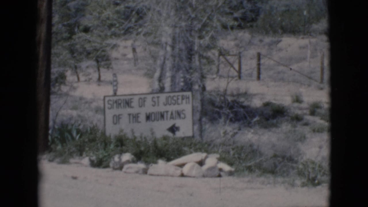 White Shrine of St Joseph of the Mountains sign beside a dirt road with shrubs and fence behind it.