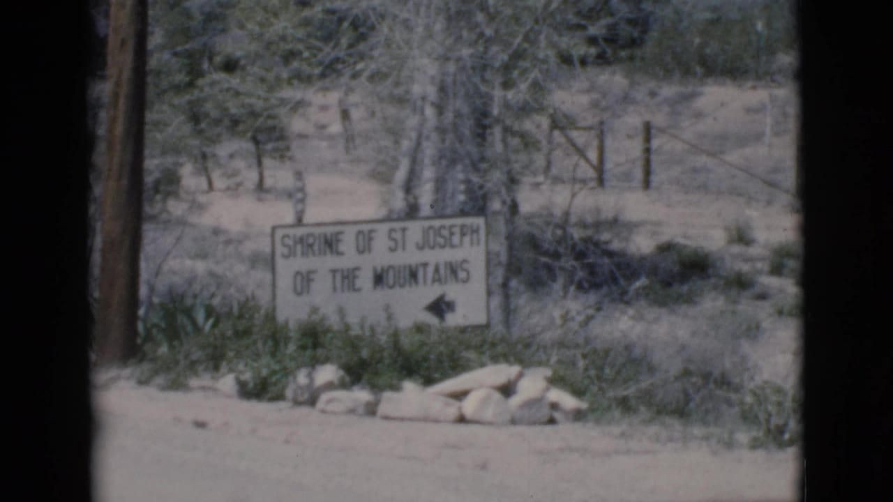 Shrine of St Joseph of the Mountains sign near brush, rocks, trees, and a wooden fence.