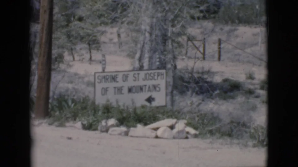 1960s 8mm frame of the Shrine of St Joseph of the Mountains sign at a rural dirt-road entrance.