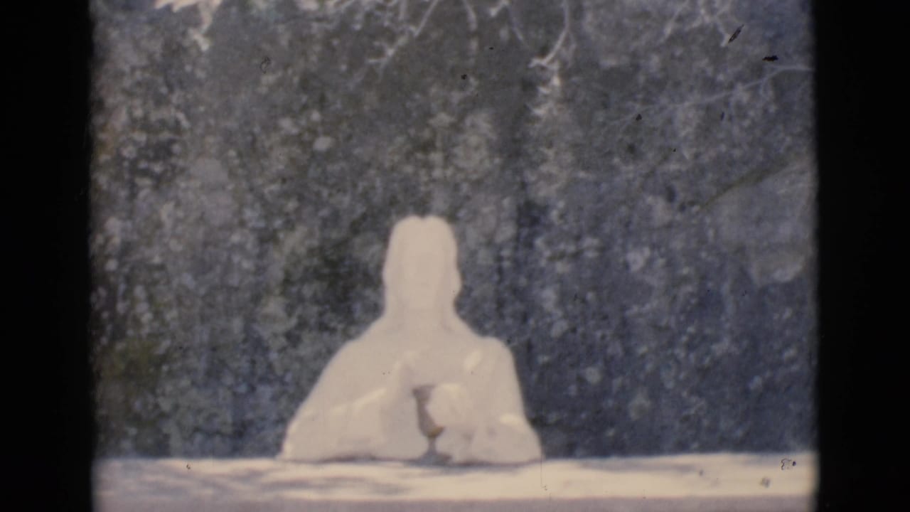 Pale humanoid clay figure seated behind a tabletop against a dark textured backdrop.