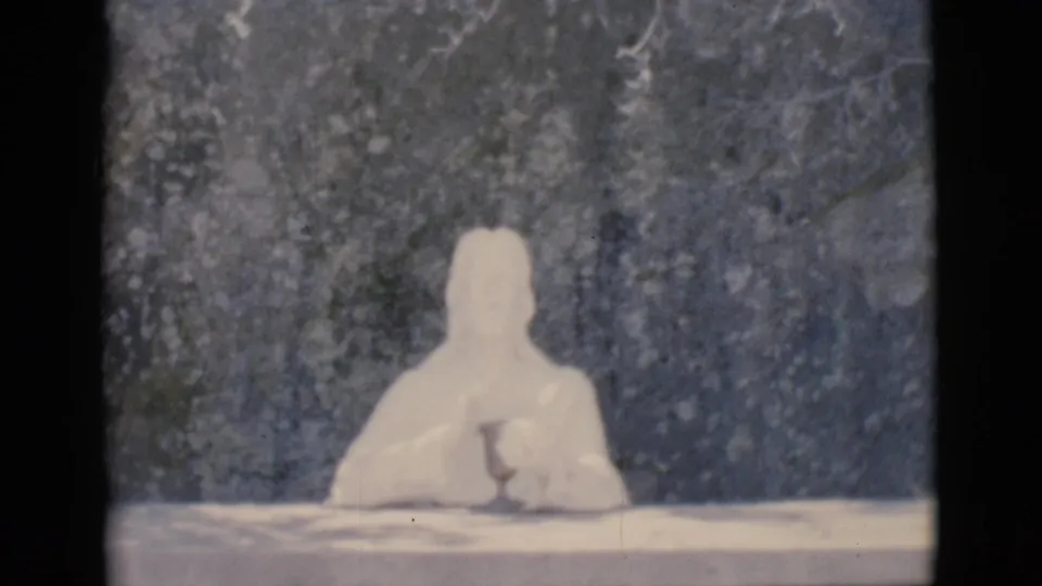 1966 Clarkdale home-movie still with a white seated humanoid figure at a tabletop, softly exposed against a mottled dark backdrop.