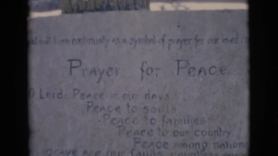 1966 home-movie frame from Clarkdale with the white statue, stone wall, and Prayer for Peace inscription in faded color.