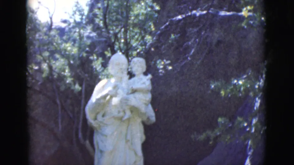 1966 Clarkdale, Arizona 8mm frame showing a white outdoor statue of a figure holding a child against trees and dark rock.