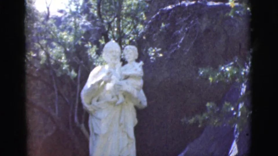 The statue of a robed figure holding a child appears centered outdoors in Clarkdale, Arizona, with trees and dark rocks behind it.