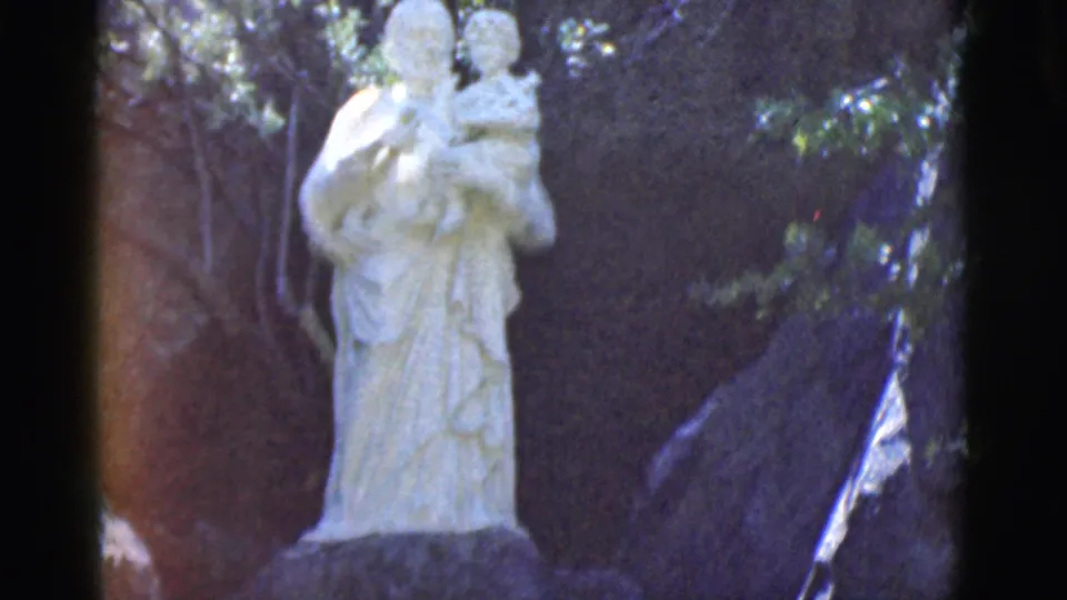 A closer 8mm frame shows the white robed statue and child figure outdoors, with foliage and shadowed rock around it.