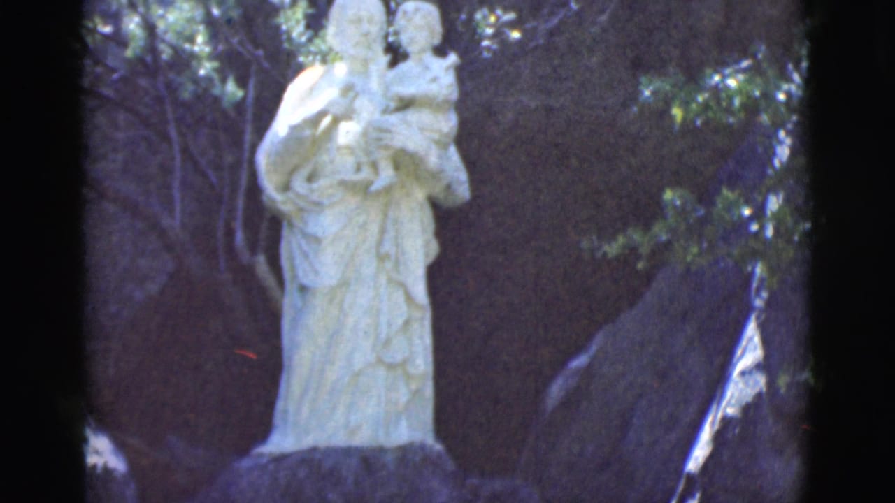 White statue of a figure holding a child among trees and rocks.