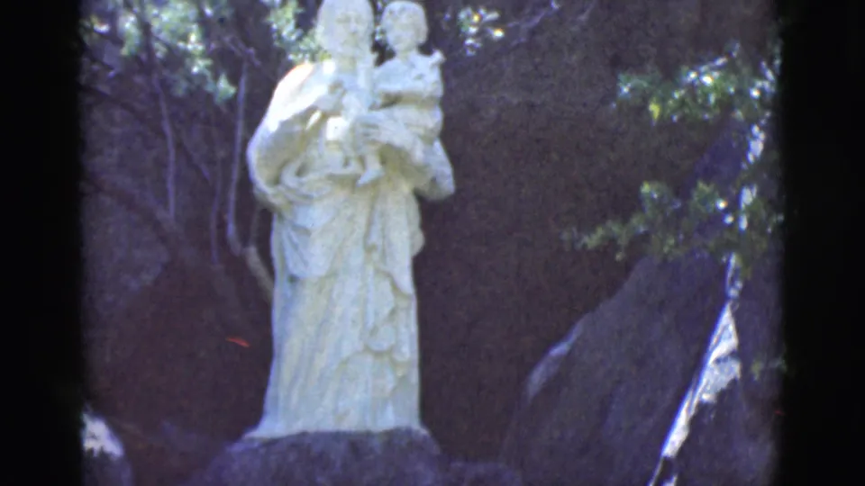 1966 Clarkdale, Arizona home-movie frame showing a pale stone statue of a figure holding a child in a shaded wooded setting.