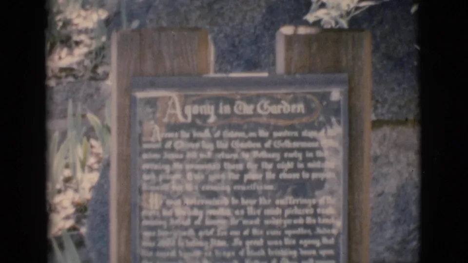 The home-movie camera holds on the Agony in the Garden plaque, with text, wood supports, stonework, and foliage visible in Clarkdale, Arizona.