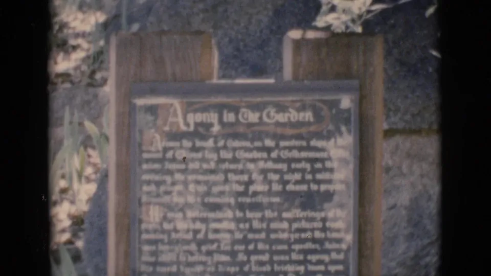 1966 Clarkdale, Arizona 8mm frame showing a wooden-framed Agony in the Garden plaque with dense text and surrounding plants.