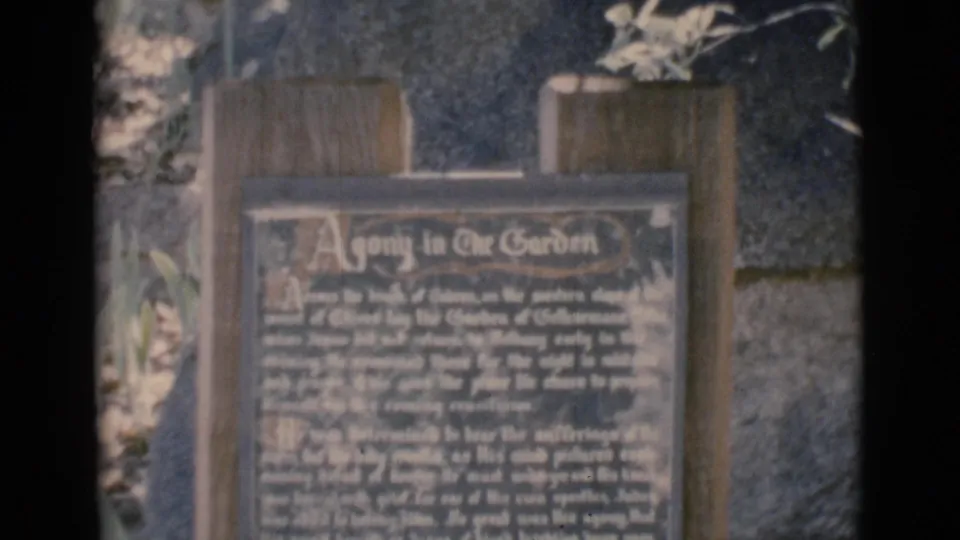 Handheld 1966 home-movie frame centered on the wooden Agony in the Garden plaque, with text softened by focus and film grain.