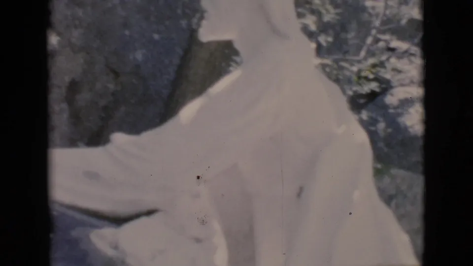 1966 Clarkdale, Arizona 8mm frame showing a close, soft-focus view of the white statue's torso, arm, rocks, and leafy background.