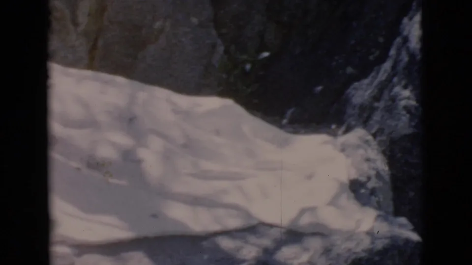1966 archival home-movie frame with white folded material across the bottom and rugged rocks behind it.