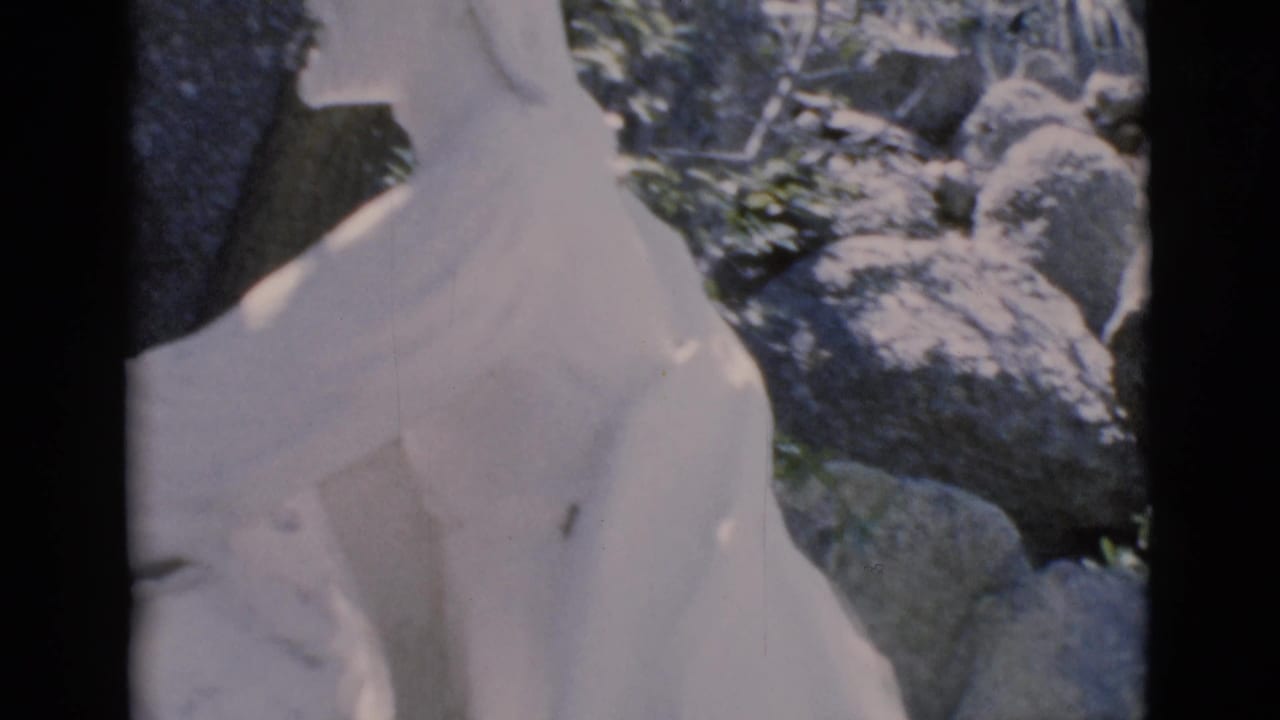 Upper section of a white robed religious statue beside gray rocks.