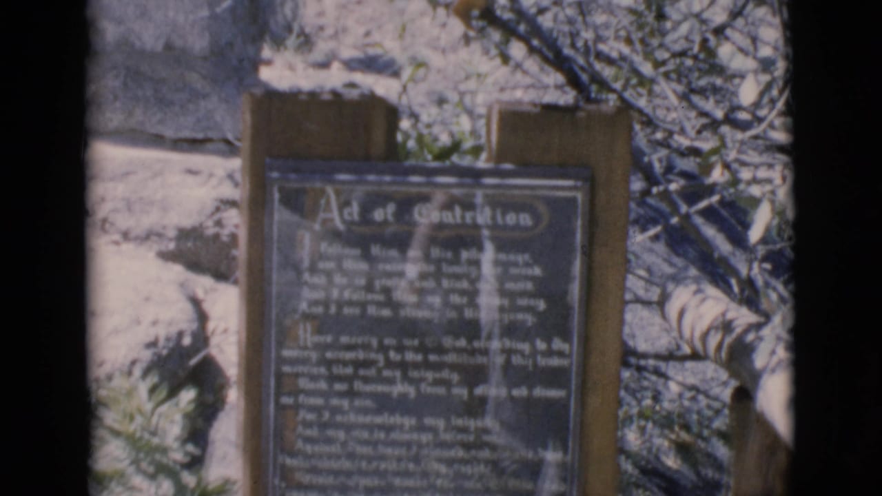 Wooden signpost with an Act of Contrition prayer plaque beside bare winter branches.