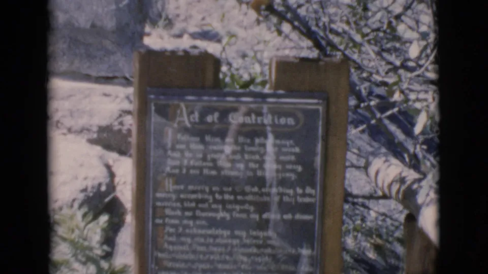1966 Clarkdale, Arizona 8mm frame showing a wooden outdoor sign with an Act of Contrition prayer plaque near bare trees.