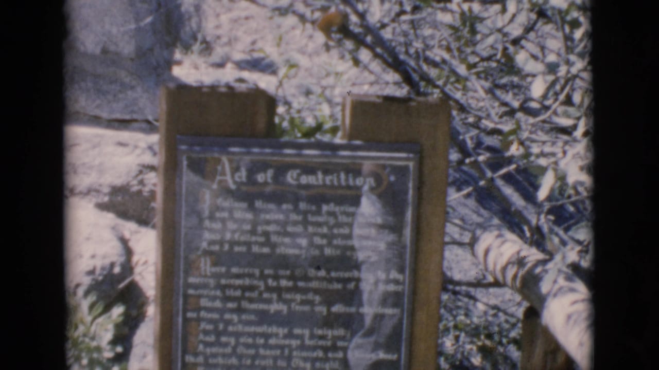 Outdoor prayer sign with white lettering, wooden posts, bare branches, and pale ground.