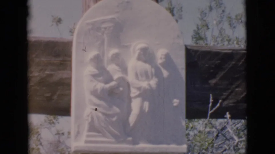 Clarkdale 1966 home-movie frame of a white carved relief depicting Jesus before figures, mounted outdoors with trees and a dark support nearby.