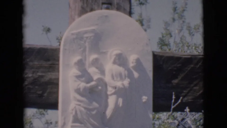 1966 Clarkdale, Arizona 8mm frame showing a white stone religious relief on a dark wooden cross with trees behind it.