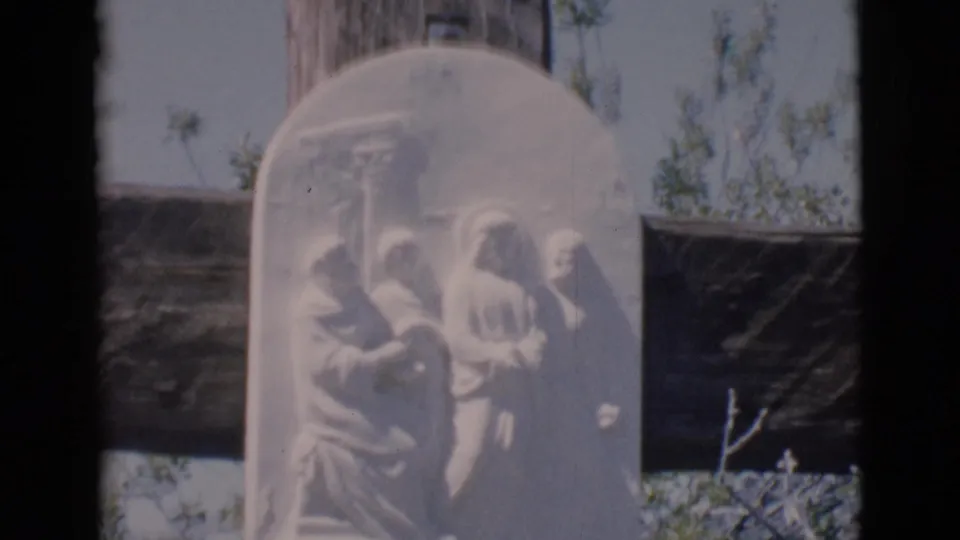 A close 8mm view of the stone relief plaque on the wooden cross, with the carved robed figures softly visible.