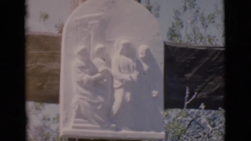 The white religious plaque nearly fills the frame, showing robed figures in relief against a dark wooden cross and trees.