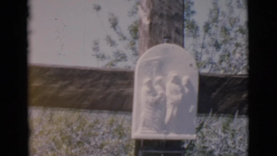 A wider 1966 home-movie frame shows the small white religious relief plaque mounted low on a large wooden cross outdoors.