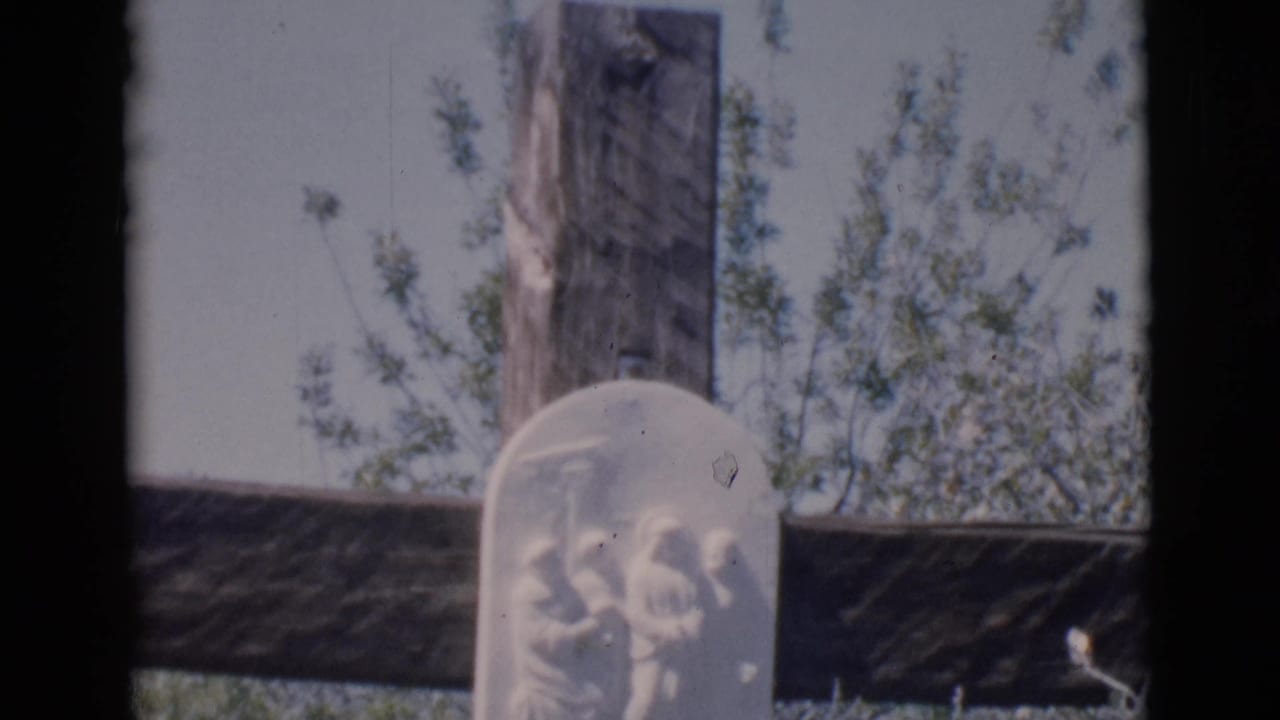 Weathered wooden cross with a pale carved religious plaque among shrubs.