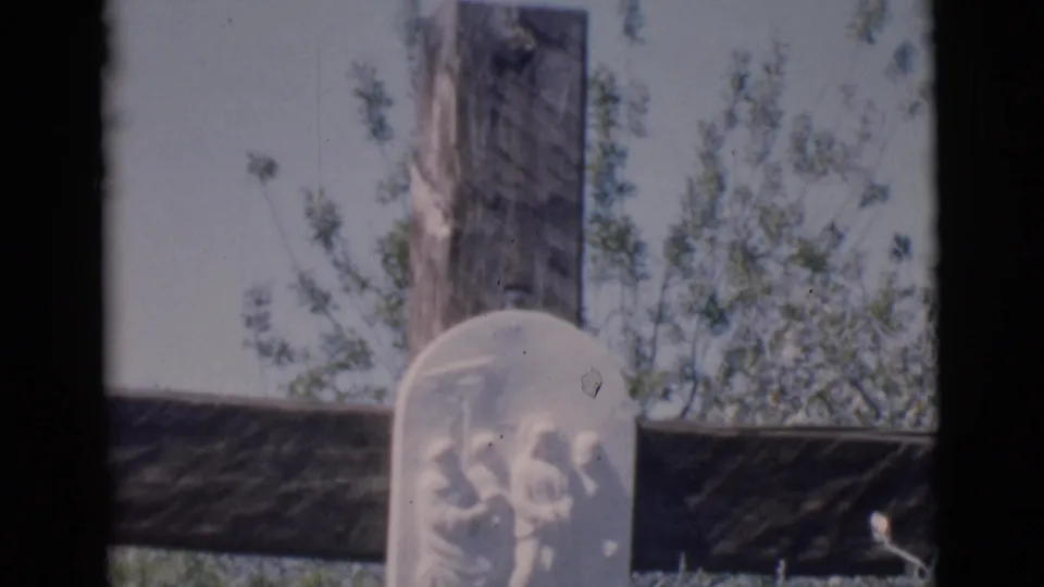 Clarkdale, Arizona 1966 8mm frame showing a wooden cross and pale carved religious memorial plaque partly surrounded by shrubbery.