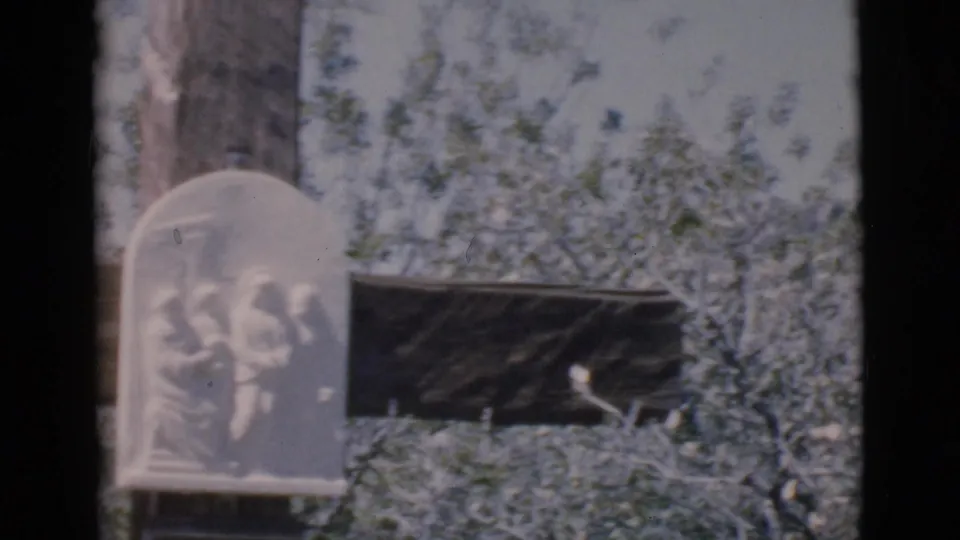 A 1966 Clarkdale home-movie frame holds on a pale carved religious plaque mounted against a weathered wooden cross outdoors.