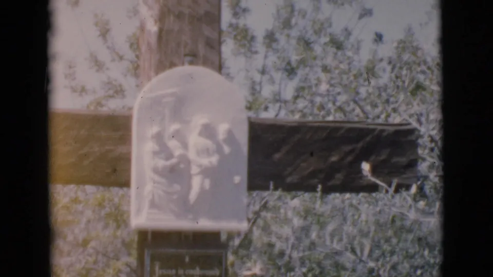 The 8mm frame shows the memorial plaque and lower inscription panel on a wooden cross in Clarkdale, Arizona, with overgrown plants nearby.