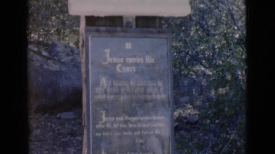 1966 Clarkdale, Arizona home-movie frame showing a close view of a religious marker reading Jesus carries His Cross among trees and rocks.