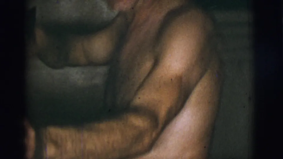 A soft, blurred 8mm frame shows the shirtless man close to bathroom fixtures, with the camera drifting during the candid moment.