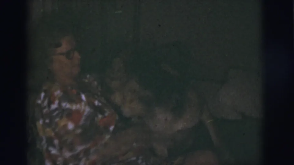 An older woman wearing glasses sits in a dim room and appears to reach toward a dog beside her on the couch.
