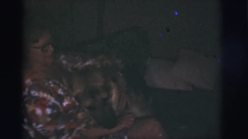 1966 Tucson home-movie frame showing a woman in a patterned dress with a German Shepherd on a sofa in a dark living room.