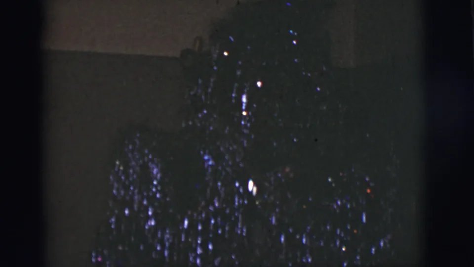 Underexposed 1966 Tucson 8mm frame showing colored lights on a dark decoration near a plain background.